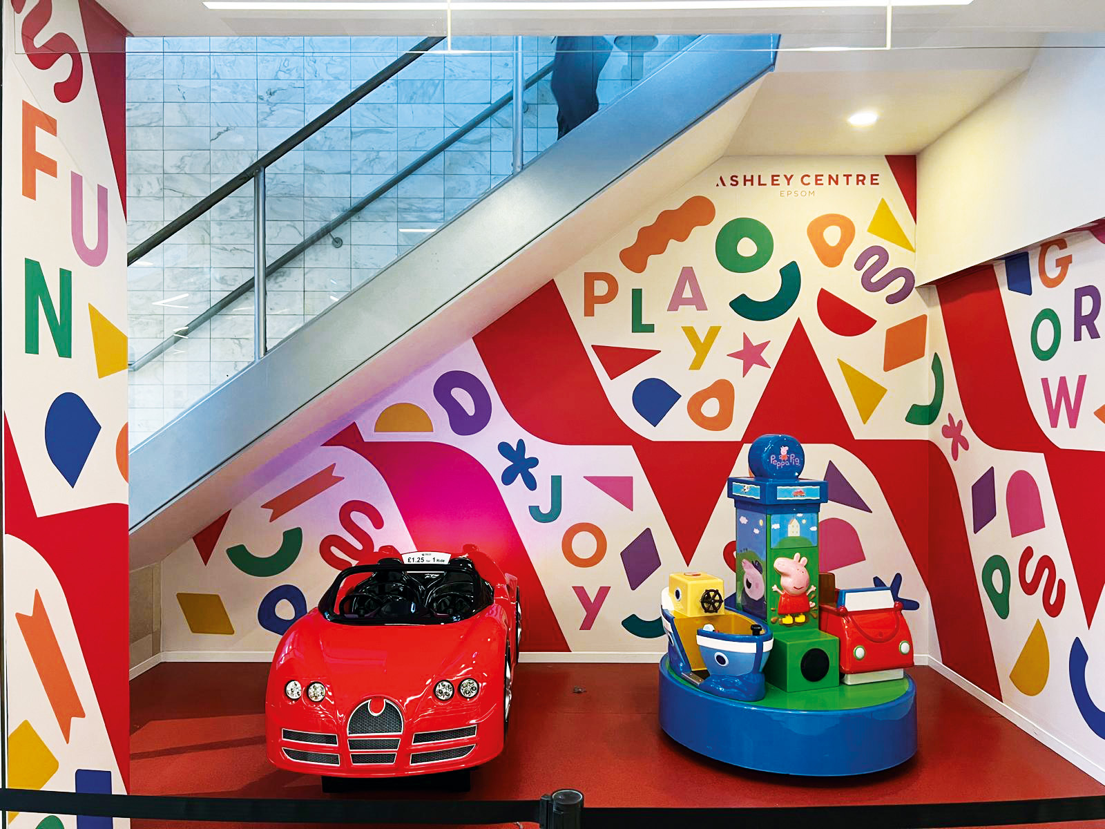 Brightly decorated childrens play area with a red toy car ride and a colorful round play structure. The background features vibrant geometric patterns and letters on the walls under a staircase.