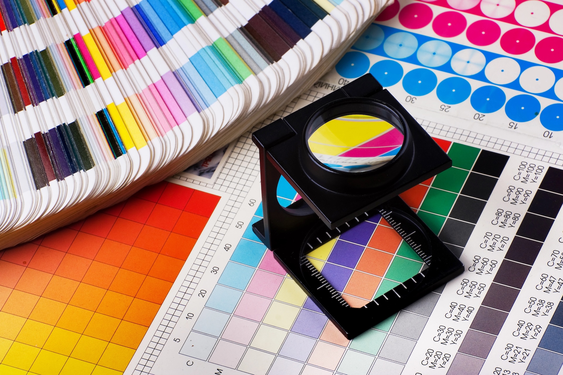 A magnifying glass sits on top of colorful printed color charts and swatches, with a fan of paper color samples nearby, illustrating graphic design and print proofing tools.
